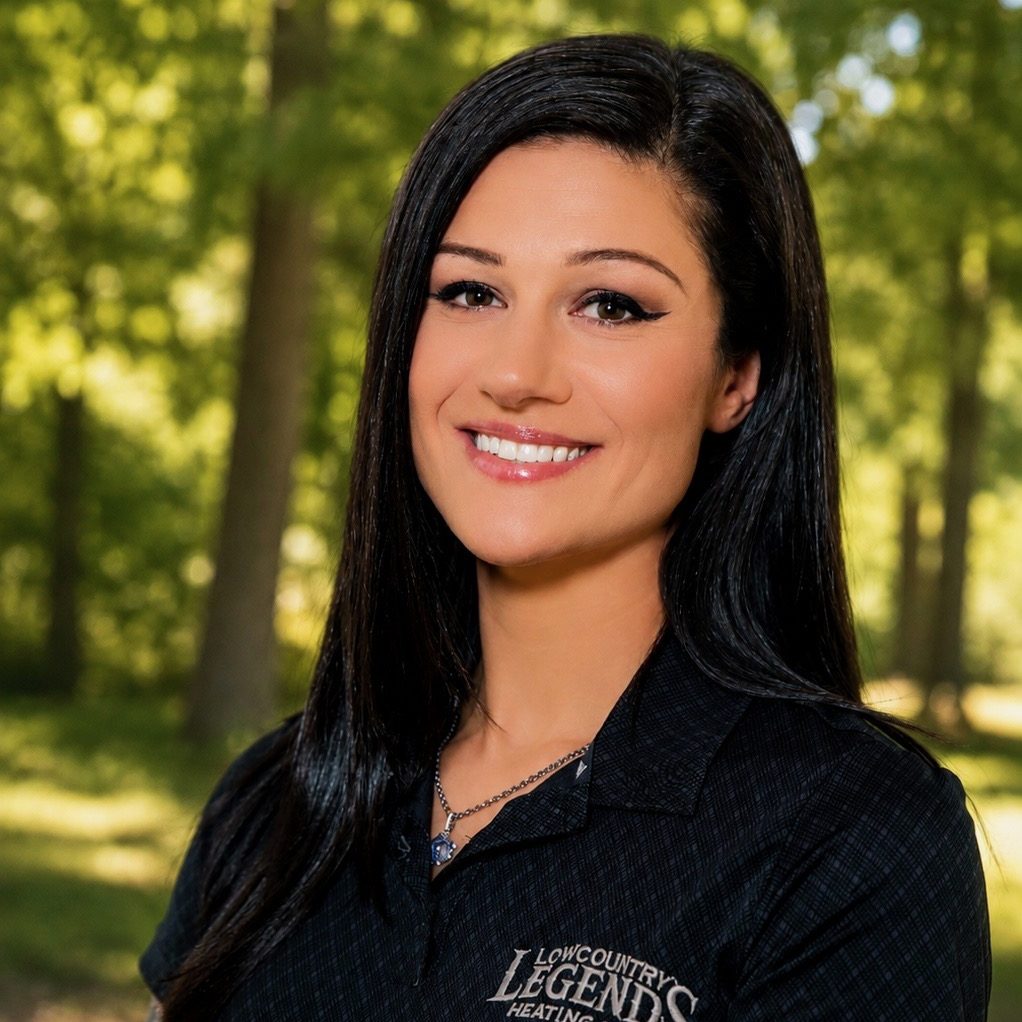 A smiling woman with long black hair wearing a black shirt with 'Lowcountry Legends Heating' logo, standing outdoors in a green forested area.